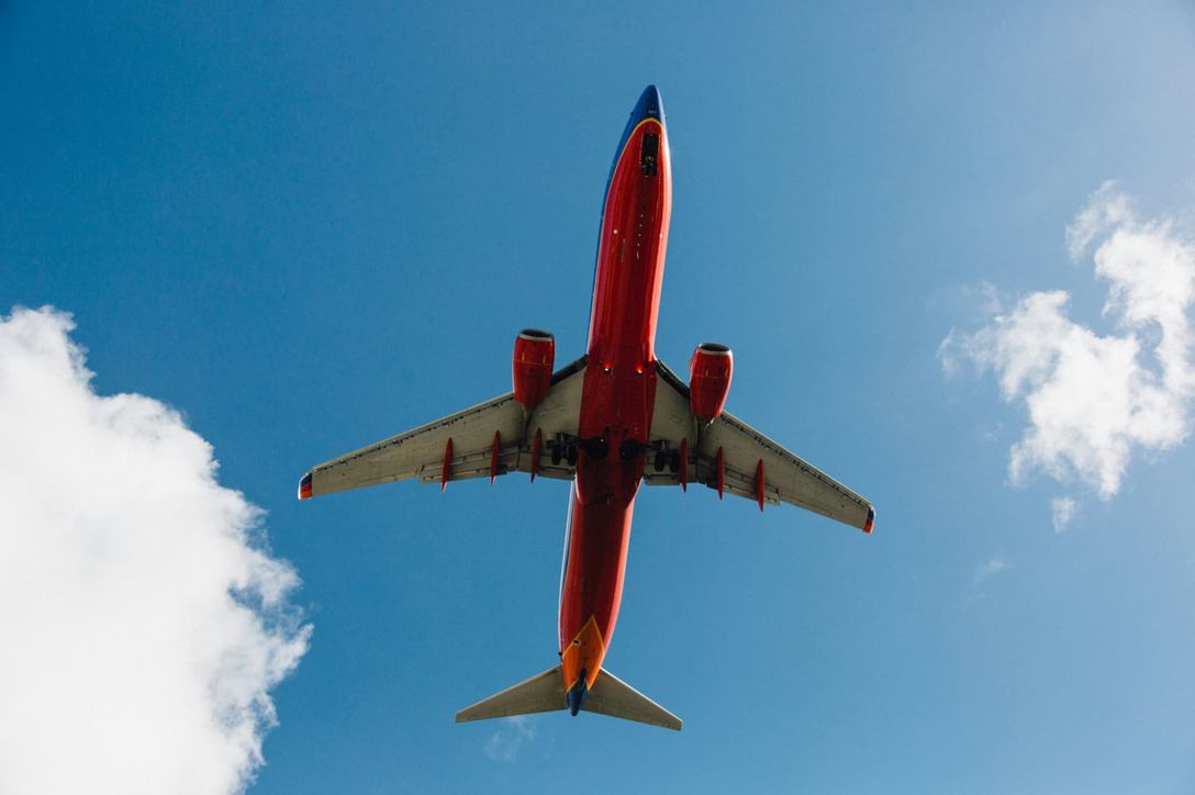 A commercial aircraft flies overhead against a blue sky with white, puffy clouds. By Kyle Glenn via Unsplash.