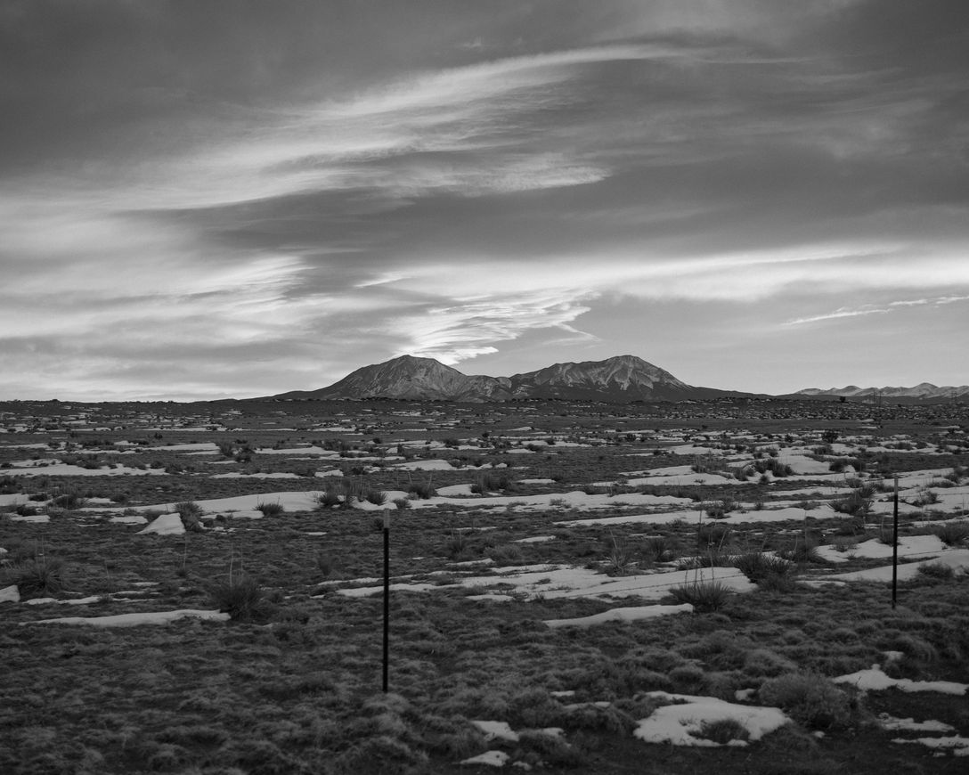 Black and white photo of sweeping plains dappled with snow and a mountain with two peaks in the distance. There's a fence in the foreground and the sky is wide and open with whispy cirrus clouds illuminated by the sun (moments from rising)